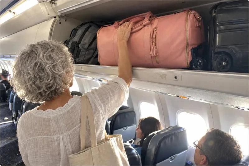 A woman lifting a pink Vohxe bag into an airplane overhead bin