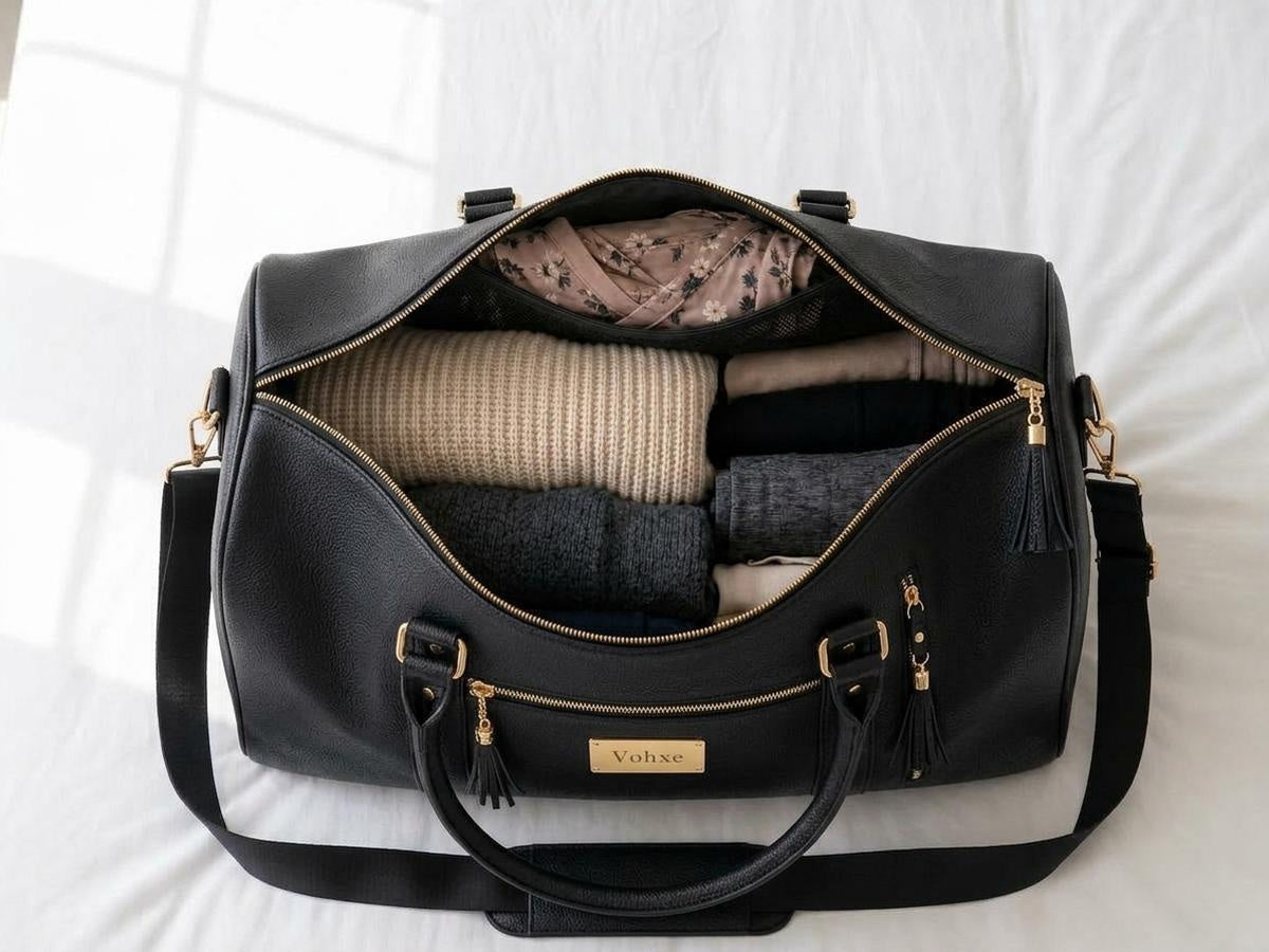 The charcoal black travel bag open on a bed, packed with folded sweaters and thermals, a wrap dress laid flat in the garment compartment