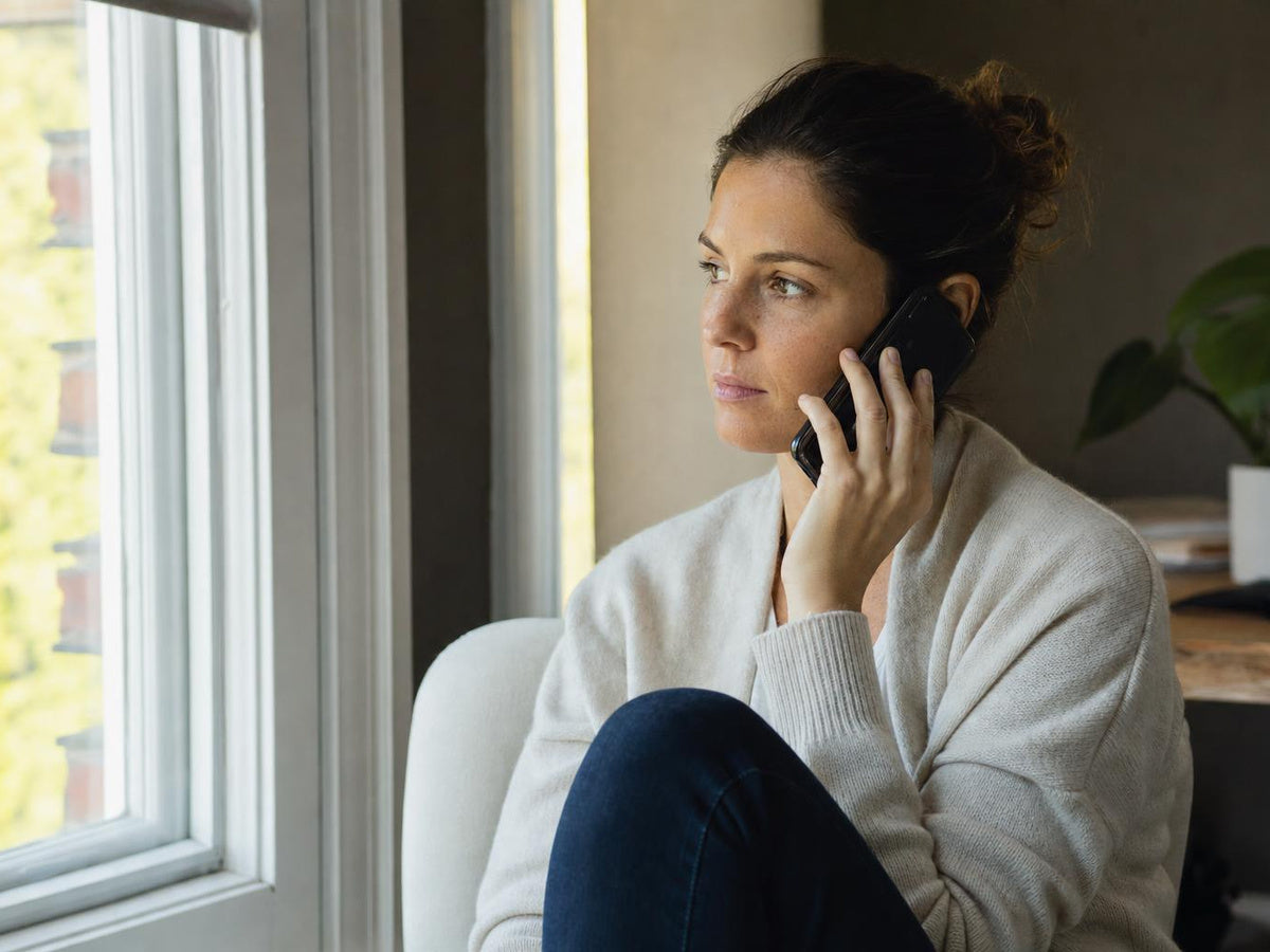 A woman sitting by a window at home, phone to her ear, reaching out to a friend for help